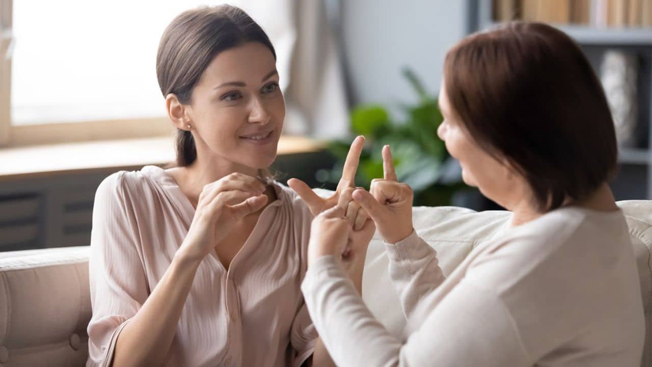 Adult daughter and middle-aged mother sit on couch at home talk using sign language, senior mum or teacher speak communicate with grownup girl child with symbols gestures, deaf-mute family concept