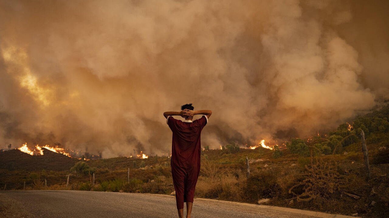 A woman looks at wildfires tearing through a forest in the region of Chefchaouen in northern Morocco on August 15, 2021. - Firefighters were battling overnight to put out two forest blazes, a forestries official said as the North African kingdom swelters in a heatwave that saw temperatures of up to 49 degrees Celsius (120 Fahrenheit) on the weekend, according to weather authorities. Morocco joins several other Mediterranean countries that have seen forest fires in recent weeks, including neighbouring Algeria where at least 90 people were killed in wildfires last week. (Photo by FADEL SENNA / AFP)