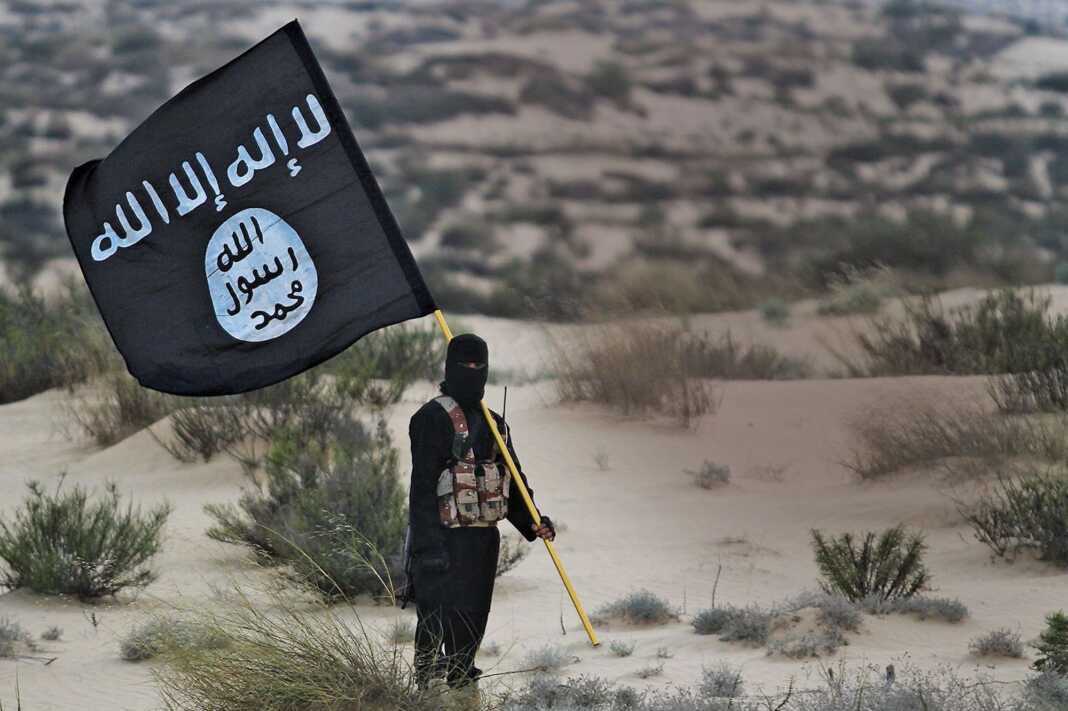 La branche de Daech en Libye revendique l’attentat contre l’ambassade d’Algérie à Tripoli Personne portant un drapeau de l'État islamique dans un paysage désertique.