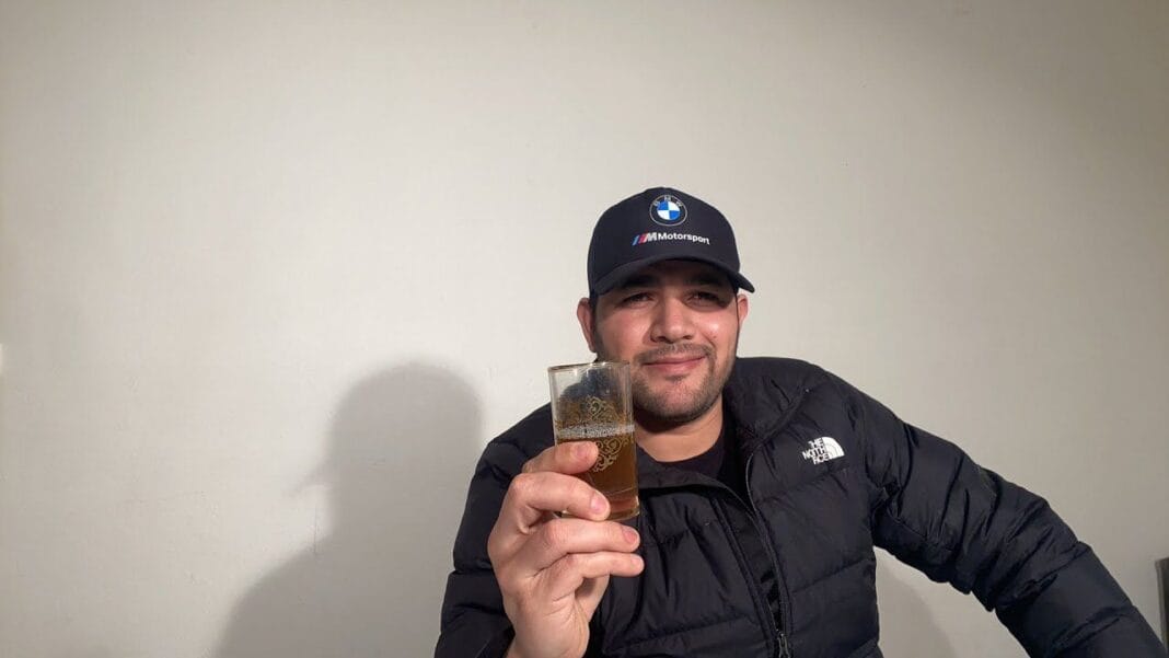 Homme avec boisson en main Homme souriant portant un chapeau et une veste noire.