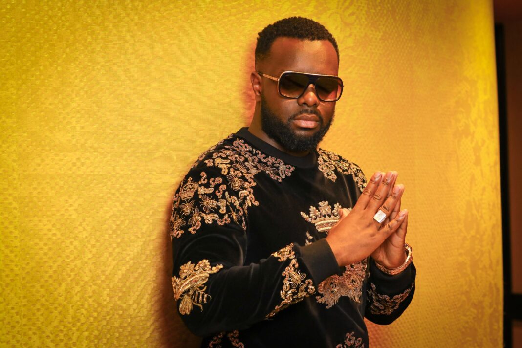 Le rappeur Gims demande aux musulmans d’arrêter de lui souhaiter une bonne année Gims portant un sweat à motifs dorés, pose avec mains jointes, fond jaune.
