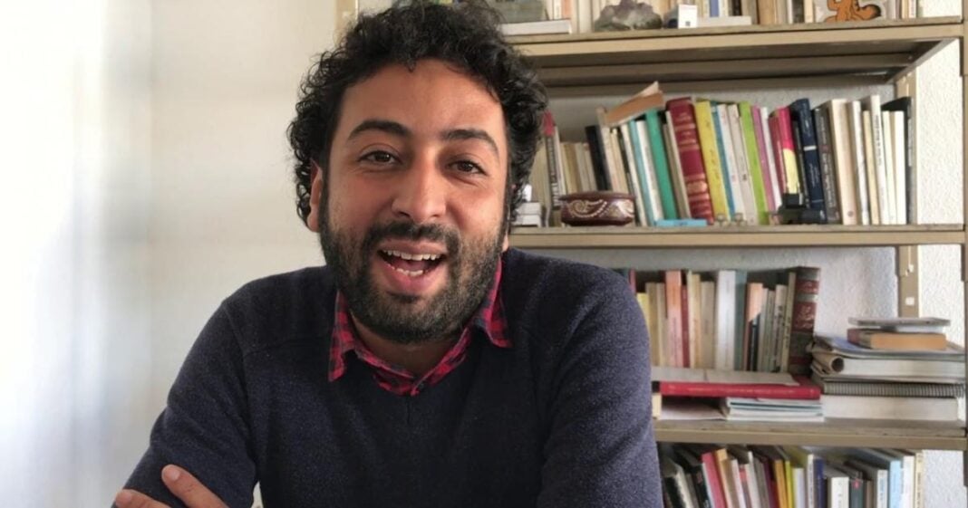 Le parquet marocain requiert 10 ans de prison contre un journaliste Homme souriant avec bibliothèque en arrière-plan.