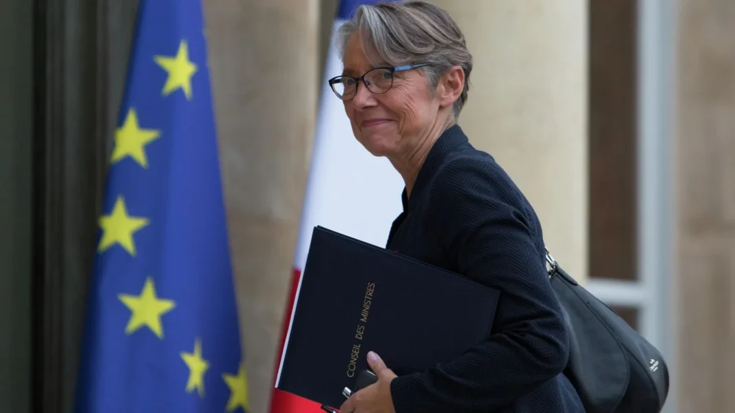 Elisabeth Borne… la première femme Premier ministre de France depuis des décennies Elisabeth Borne avec un dossier officiel, souriante, devant un drapeau européen.