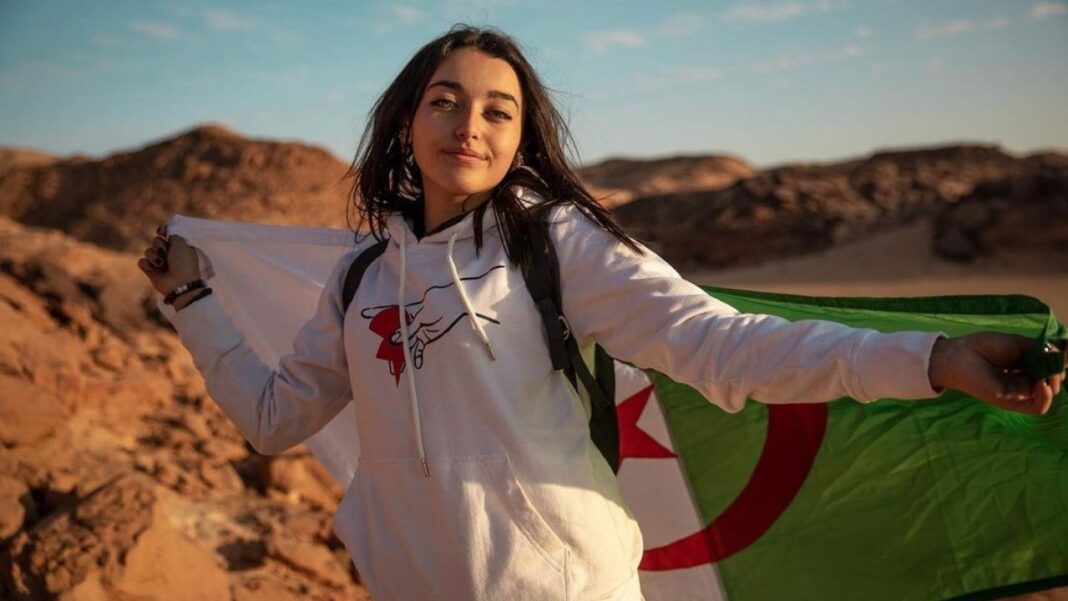 Affaire Future Gate en Algérie: accusée de participation à des escroqueries, Ines Abdelli écope de… Femme souriante tenant un drapeau algérien dans un paysage désertique.
