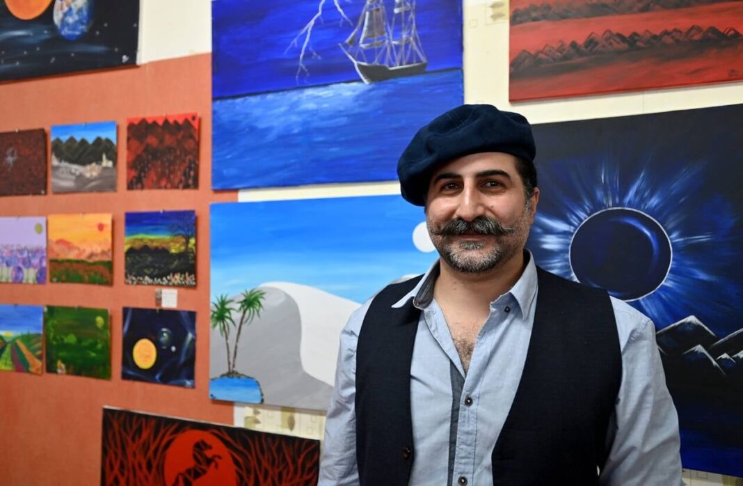 Exclu : Le fils de Ben Laden expose ses toiles à Paris Homme souriant dans une galerie d'art avec peintures colorées en arrière-plan.