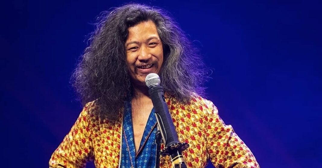 Qu’est ce qu’un algérien selon Bun Hay Mean ? Bun Hay Mean avec cheveux bouclés et sourire, parlant dans un micro.