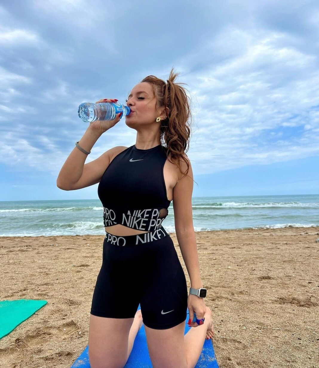 Femme sportive buvant de l'eau sur la plage par temps nuageux.