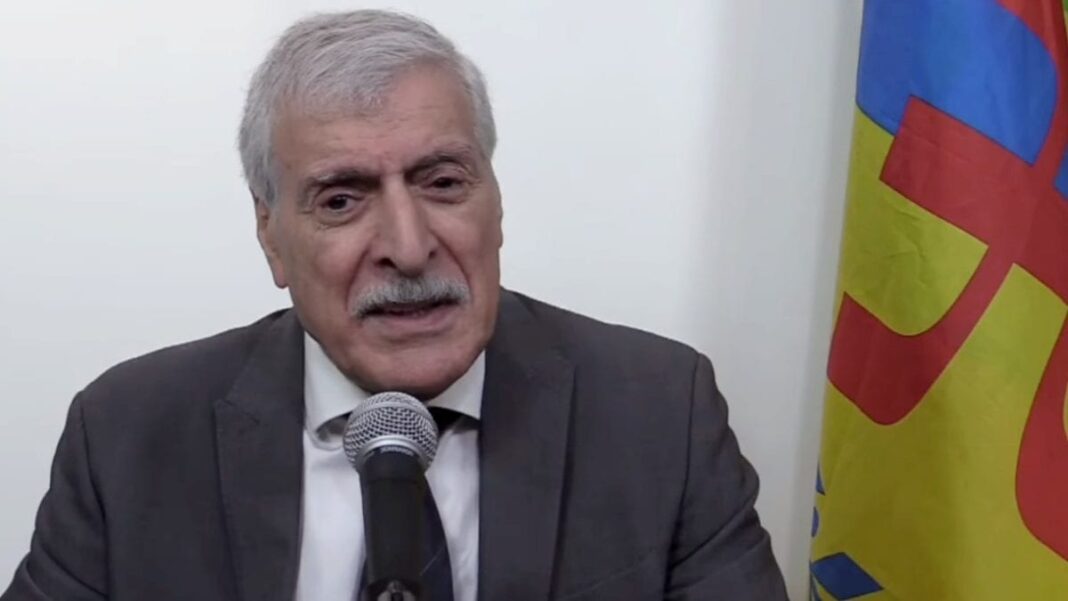 Justice Algérie : Ferhat Mehenni condamné par coutumace à la perpétuité Ferhat Mehenni parlant en public avec drapeau kabyle en arrière-plan.