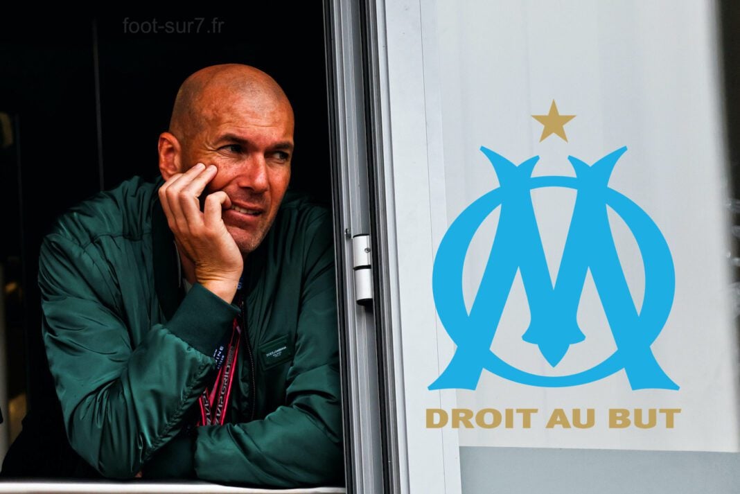 Zinedine Zidane à l’OM ? Samir Nasri fait de croustillantes révélations Zinedine Zidane regardant pensivement par une fenêtre avec logo OM.