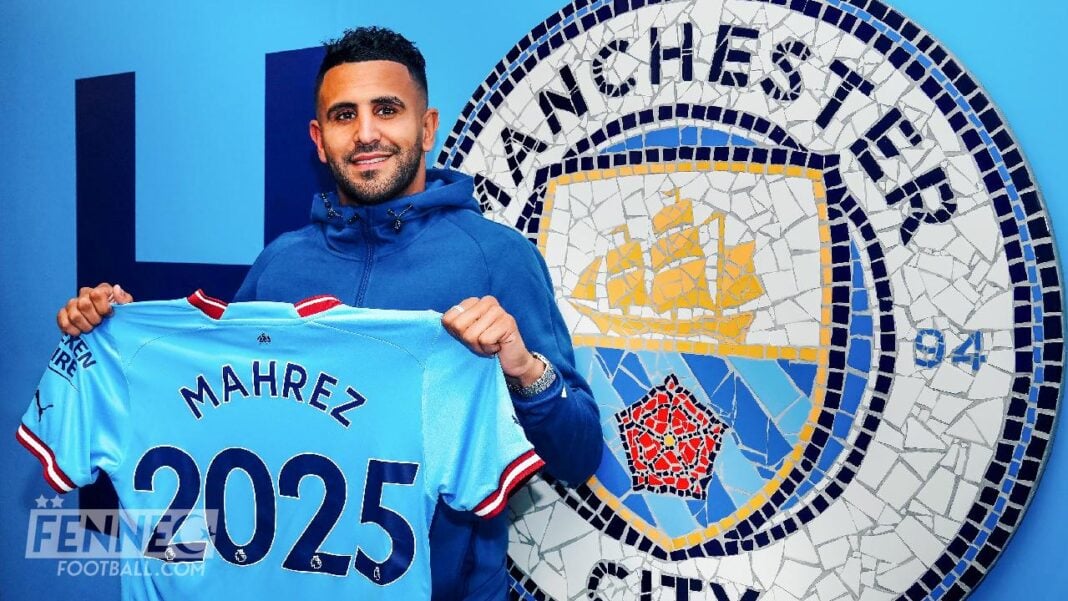 Riyad Mahrez tenant un maillot de Manchester City en face du logo du club.