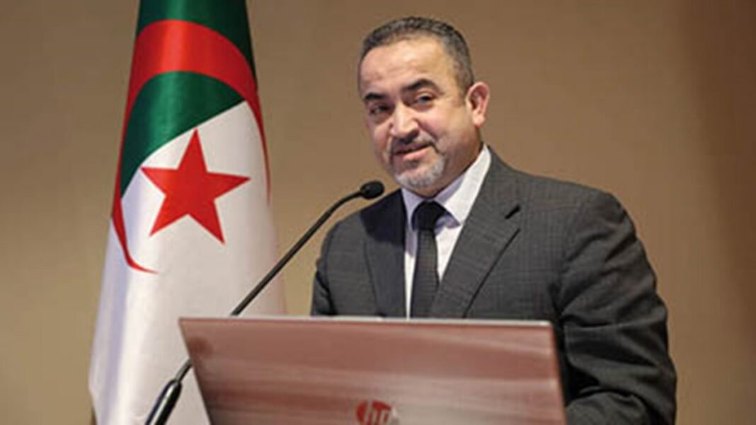 Coup de Tonnerre ! A propos de l’affaire Berraki Arzeki, les épouses de l’homme font de terribles aveux ! Homme en costume devant drapeau algérien lors d'une intervention.
