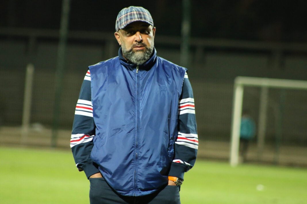 Entraîneur de football sur le terrain Entraîneur de football en vestiaire, regard sérieux, stade en arrière-plan.