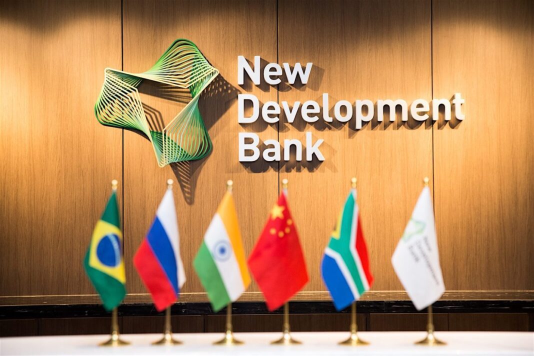 Officiel ! La NDB, la banque des BRICS, dit oui à l’adhésion de l’Algérie. Logo de la NDB avec drapeaux des pays BRICS en premier plan.