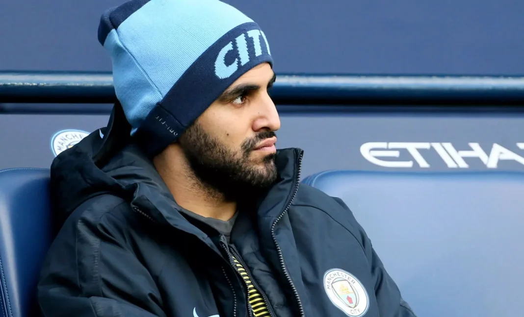 Mahrez vient d’être viré par City ? Mahrez assis, portant un bonnet et une veste de Manchester City.