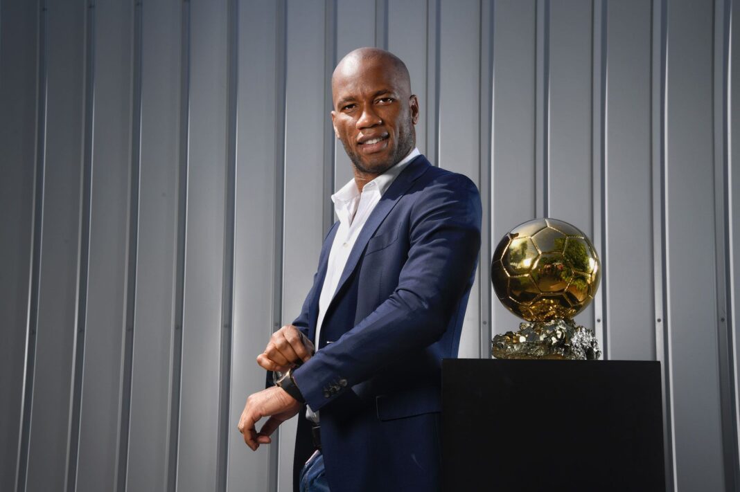 Didier Drogba ne connaît pas Cristiano et Messi : voici ses trois plus grands joueurs de l’histoire Didier Drogba en costume avec un ballon dor en arrière-plan.