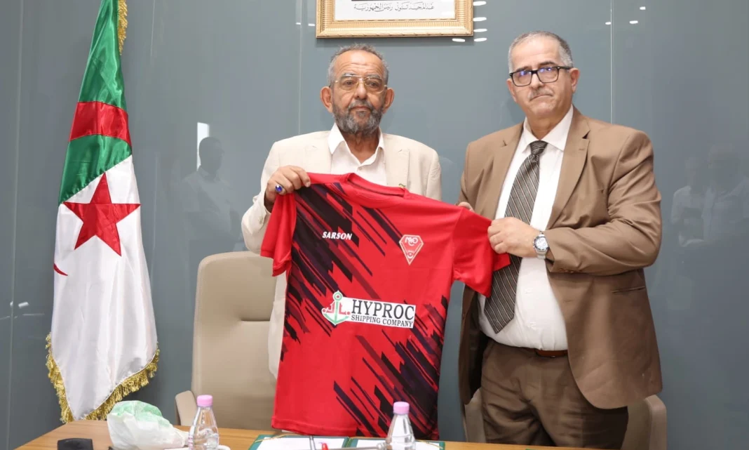 C’est officiel! Le MC Oran est racheté par Hyproc Deux hommes en costume présentent un maillot du MC Oran lors d'une cérémonie officielle.