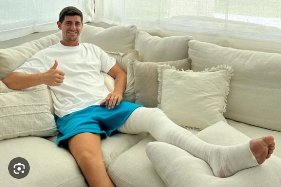 Real Madrid : Thibaut Courtois sort d’une opération réussie Jeune homme souriant avec jambe en plâtre sur canapé blanc.