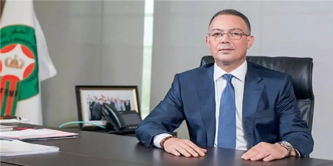 Le Maroc organisera la coupe du monde féminine 2031 ! Homme en costume dans un bureau avec drapeau marocain en arrière-plan.