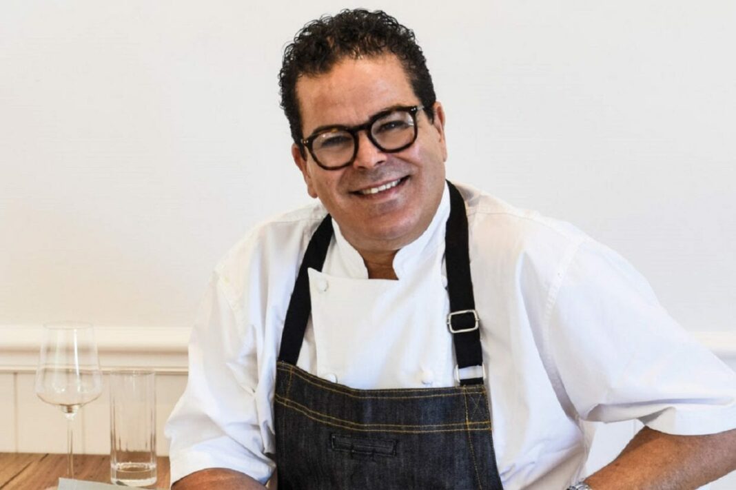 Diaspora Algérienne : Les révélations étonnantes du chef renommé Pierre Khodja Chef Pierre Khodja souriant en tenue de cuisine avec un verre à côté.