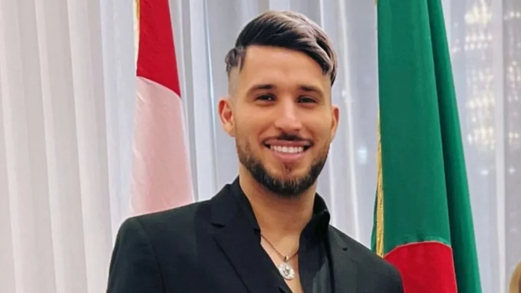Fayçal El-Mignon souriant, portant un costume noir, devant un drapeau algérien et un drapeau étrangé.