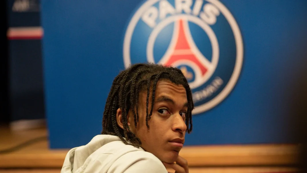 Ethan Mbappe, un talent brut à polir Jeune joueur de football devant le logo du PSG.