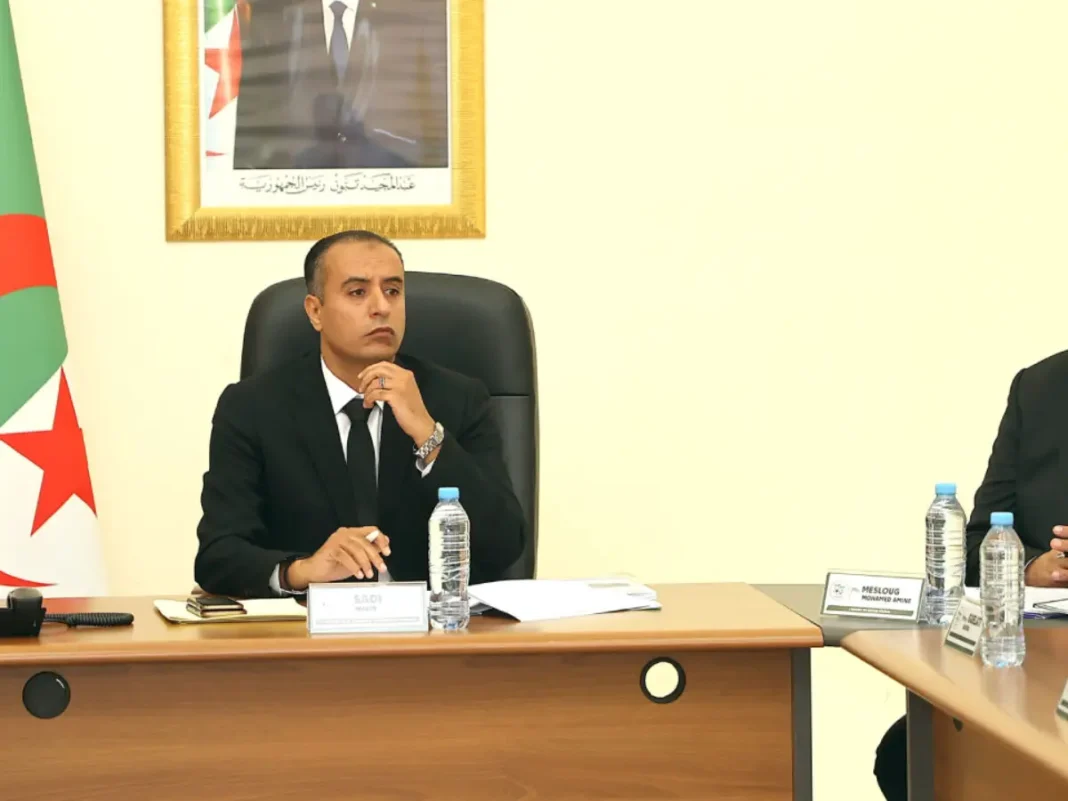 Walid Sadi lors d'une réunion officielle Walid Sadi en réunion, portant un costume noir, dans un bureau avec drapeau algérien en arrière-plan.