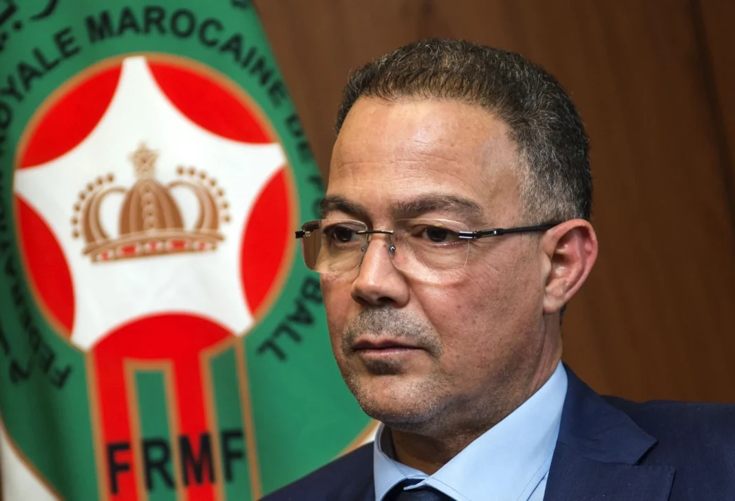Scandale à la CAN : Un arbitre marocain accusé de corruption Fouzi Lekjaa en costume avec le logo du Maroc en arrière-plan.
