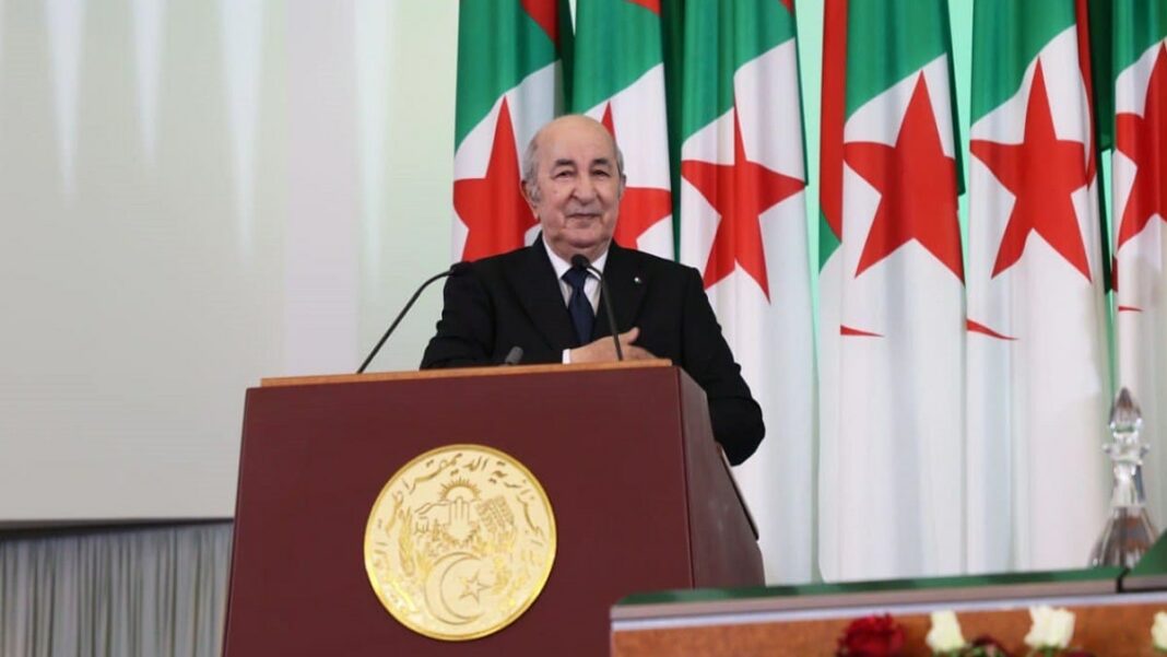 Journée internationale des travailleurs : Tebboune honore deux héros nationaux Président Tebboune parlant à un podium avec drapeaux algériens en arrière-plan.