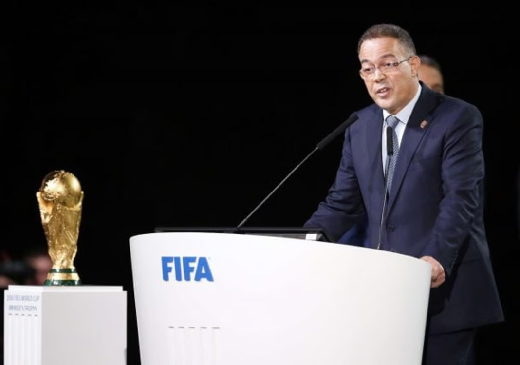 Homme en costume parlant à une conférence FIFA avec trophée à côté.