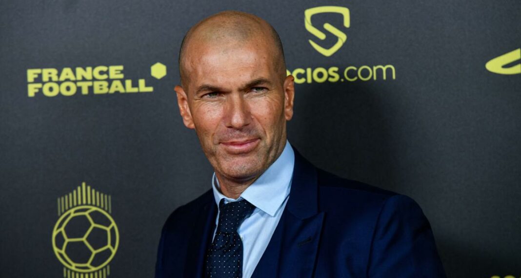 Zinedine Zidane lors d'une conférence, visage concentré, fond de logo football et médias.