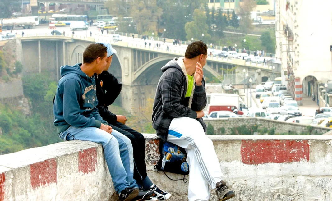 Indice du Bonheur 2024 : Algérie, Déclin du Bonheur et Fractures Générationnelles Jeunes hommes assis sur un mur avec un pont en arrière-plan à Alger.