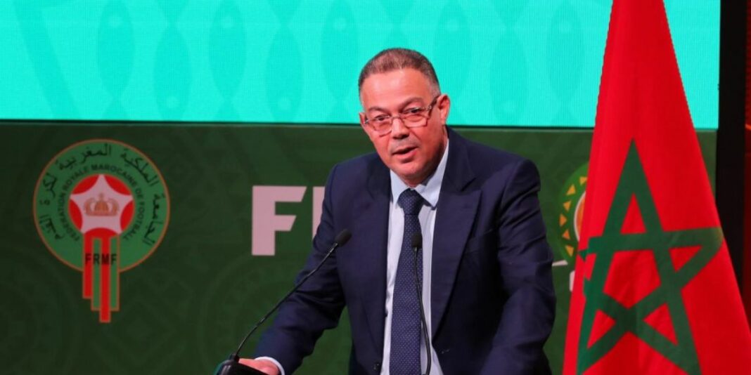 Maroc : Fouzi Lekjaa brave la CAF et annonce les dates potentielles de la CAN 2025 ! Fouzi Lekjaa parle lors d'une conférence avec drapeau marocain en arrière-plan.