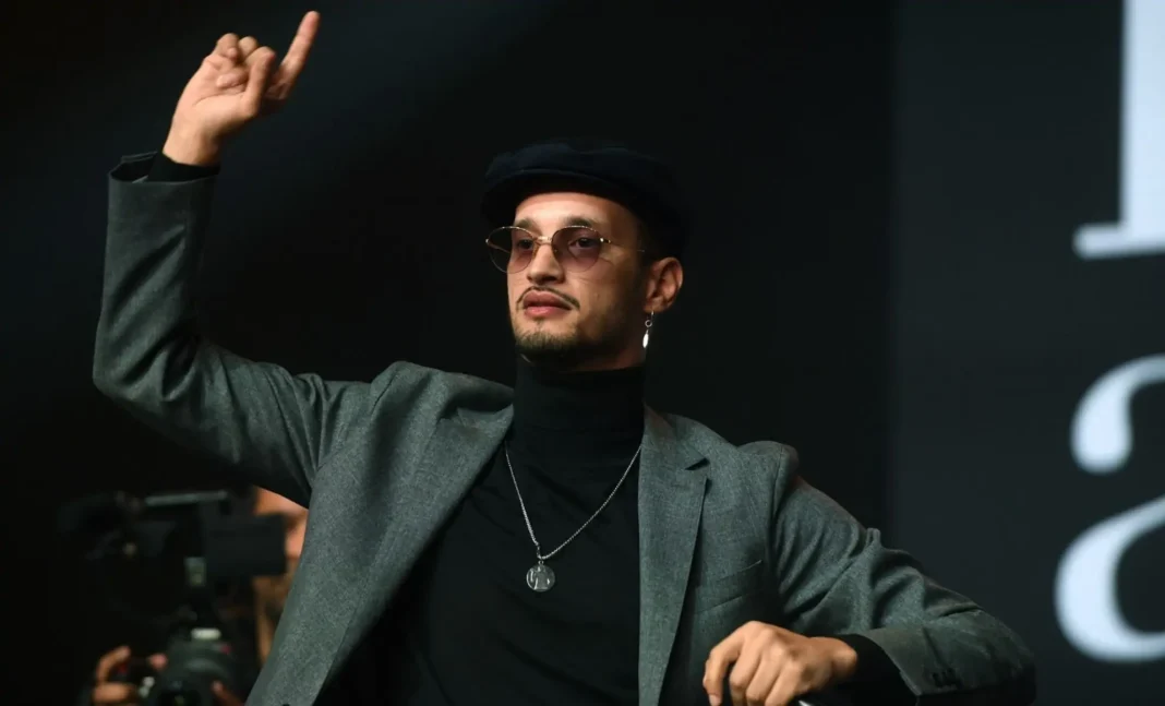 « La musique est haram » : Soolking déclenche une grosse polémique Soolking en costume avec lunettes, levant le doigt lors d'une intervention.