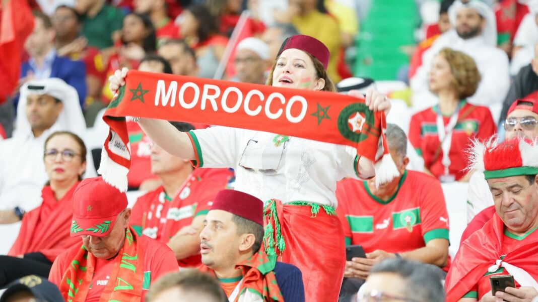 CAN 2025 : Le Maroc au Bord du Gouffre !…. une Course Contre la Montre à Haut Risque ! Supporters marocains en tenue traditionnelle et avec drapeau lors d'un match de football.