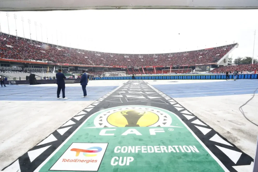 Vue du terrain de football avec logo de la CAF et stade rempli en arrière-plan.