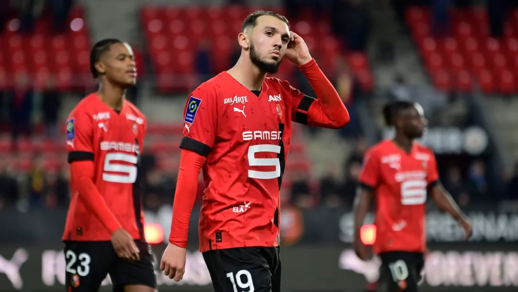 Amine Gouiri reste à Rennes ? Le joueur se lâche Amine Gouiri en tenue de football rouge, pensif sur le terrain de Rennes.