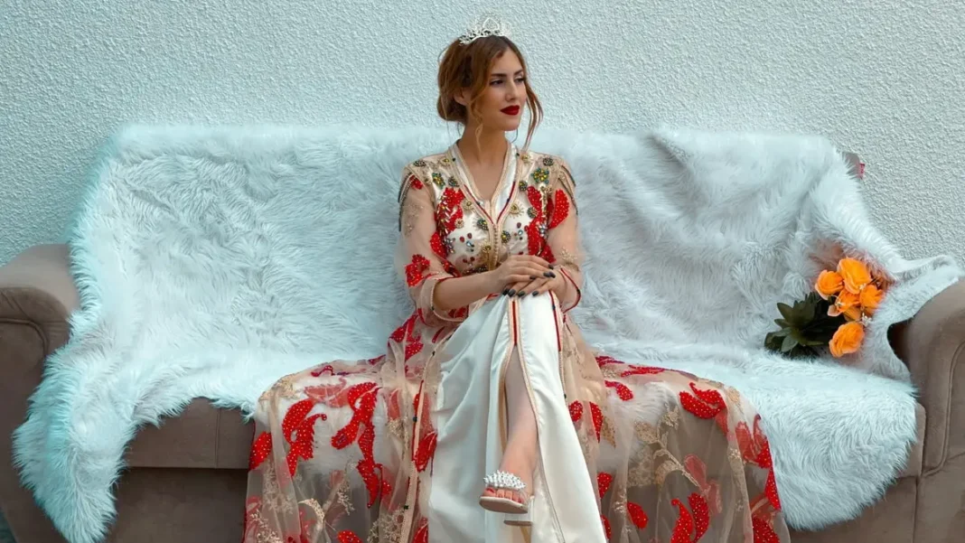 Caftan : Nouvelle offensive du Maroc pour s’approprier un patrimoine maghrébin partagé Femme portant un caftan marocain traditionnel avec broderies rouges et or, assise sur un canapé.