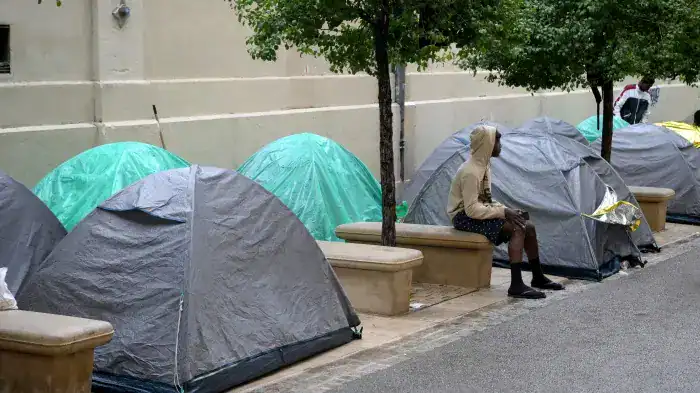 Tentes de migrants installées sur le trottoir avec une personne assise à proximité.