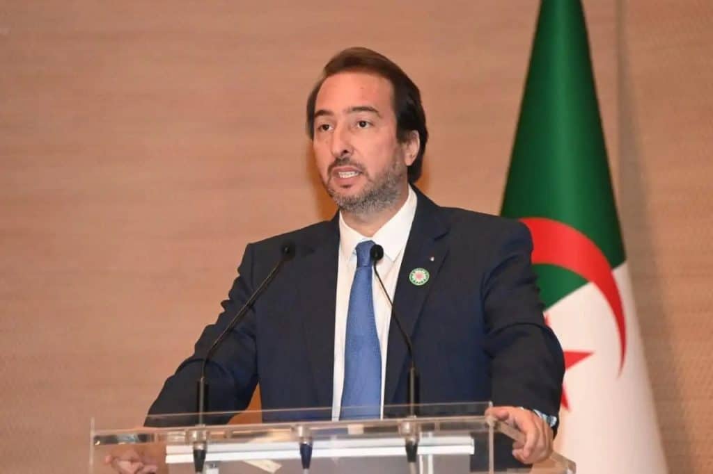 Homme politique parlant lors d'une conférence avec drapeau algérien en arrière-plan.