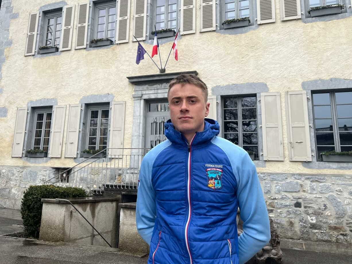 Il perd sa naturalisation à cause de son salaire trop bas Jeune homme en veste bleue devant une mairie avec drapeaux français et européen.