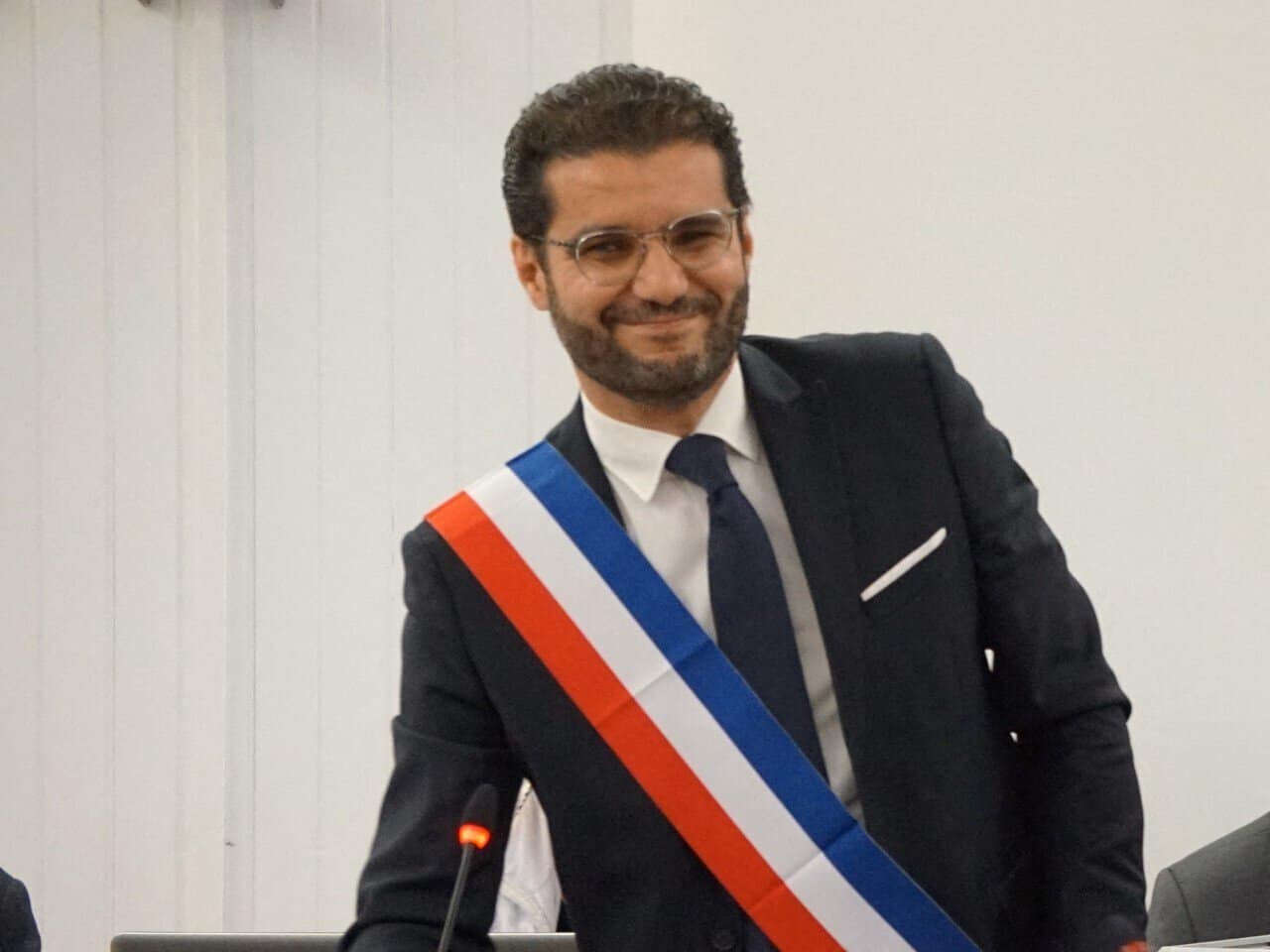 Un Franco-Algérien défie les attentes et devient maire ! Homme en costume avec écharpe tricolore, sourire confiant.