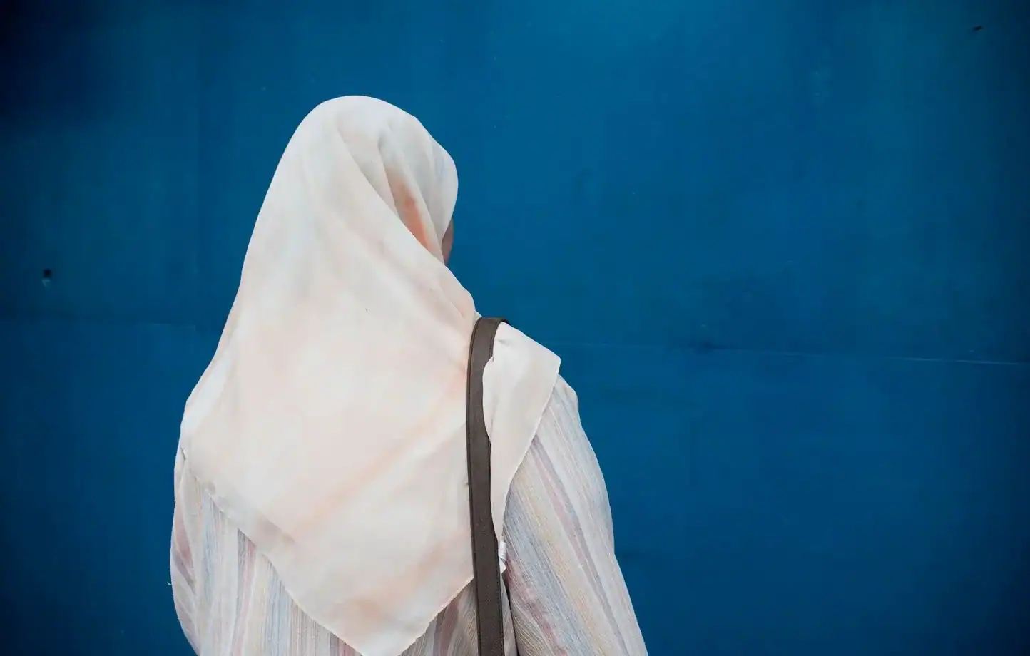 France : quand le voile coûte leur emploi aux salariées Femme avec hijab blanc vue de dos devant un mur bleu.