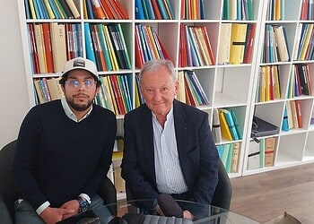 Deux hommes assis à une table en bibliothèque, livres en arrière-plan.